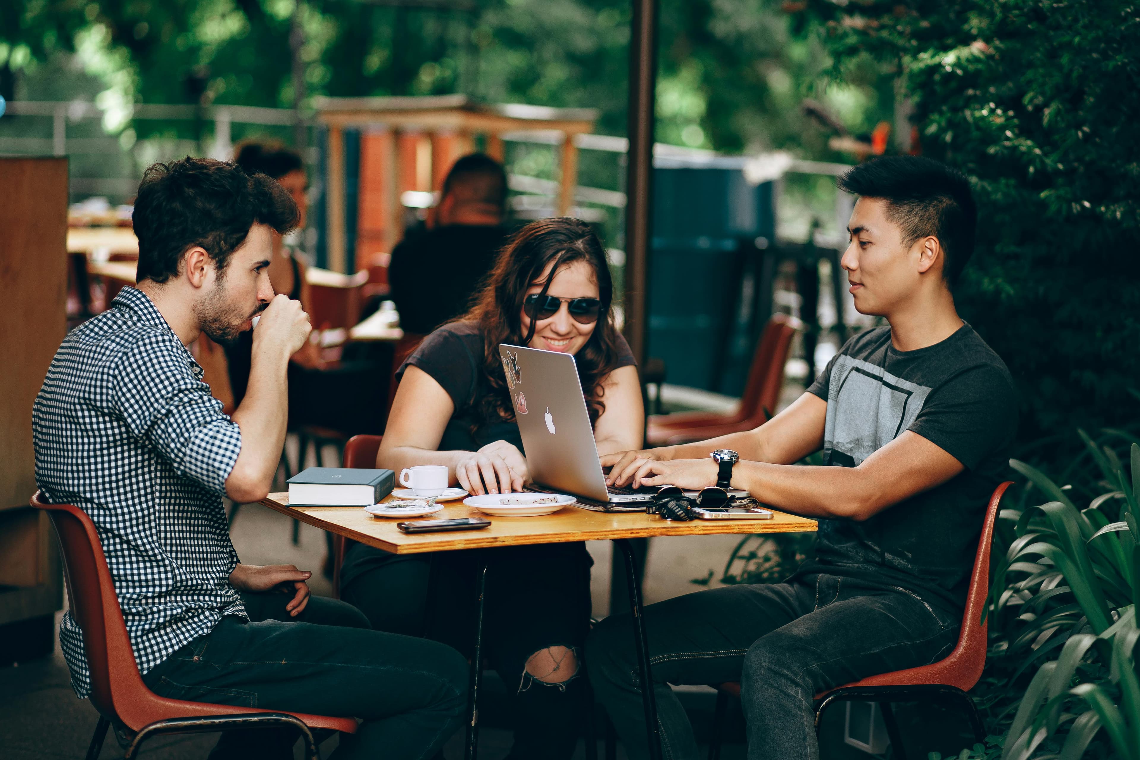 Three friends chatting at an outdoor cafe table with a laptop